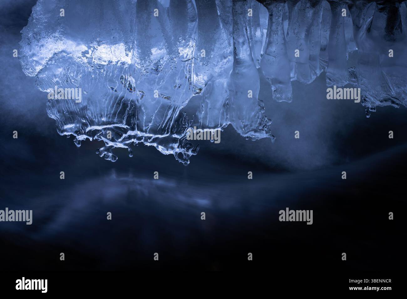 Skulpturale Eisformationen hängen über einem Fluss aus dunklem, beweglichem Wasser, beleuchtet in kühlen Blautönen. Stockfoto