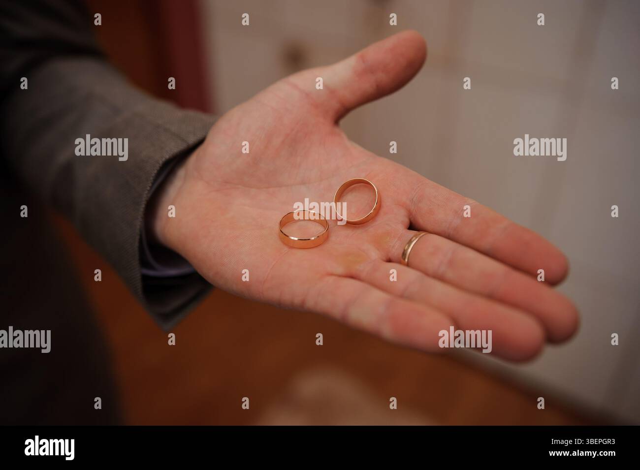 Eine Nahaufnahme zeigt die Hand eines Mannes, der zwei einfache goldene Eheringe hält, die Ehe, Liebe und Bindung symbolisieren, gegen ein verschwommenes, warmes ich Stockfoto