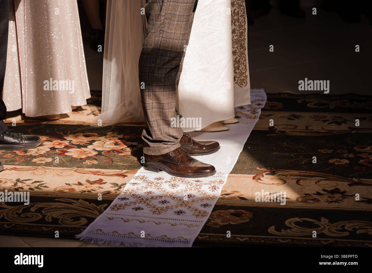 Eine warm beleuchtete Indoor-Szene zeigt eine Hochzeitszeremonie mit einem Bräutigam, der auf einem traditionellen Teppichläufer während des Glasbruchs steht Stockfoto Eine warm beleuchtete Indoor-Szene zeigt eine Hochzeitszeremonie mit einem Bräutigam, der auf einem traditionellen Teppichläufer während des Glasbruchs steht Stockfoto
