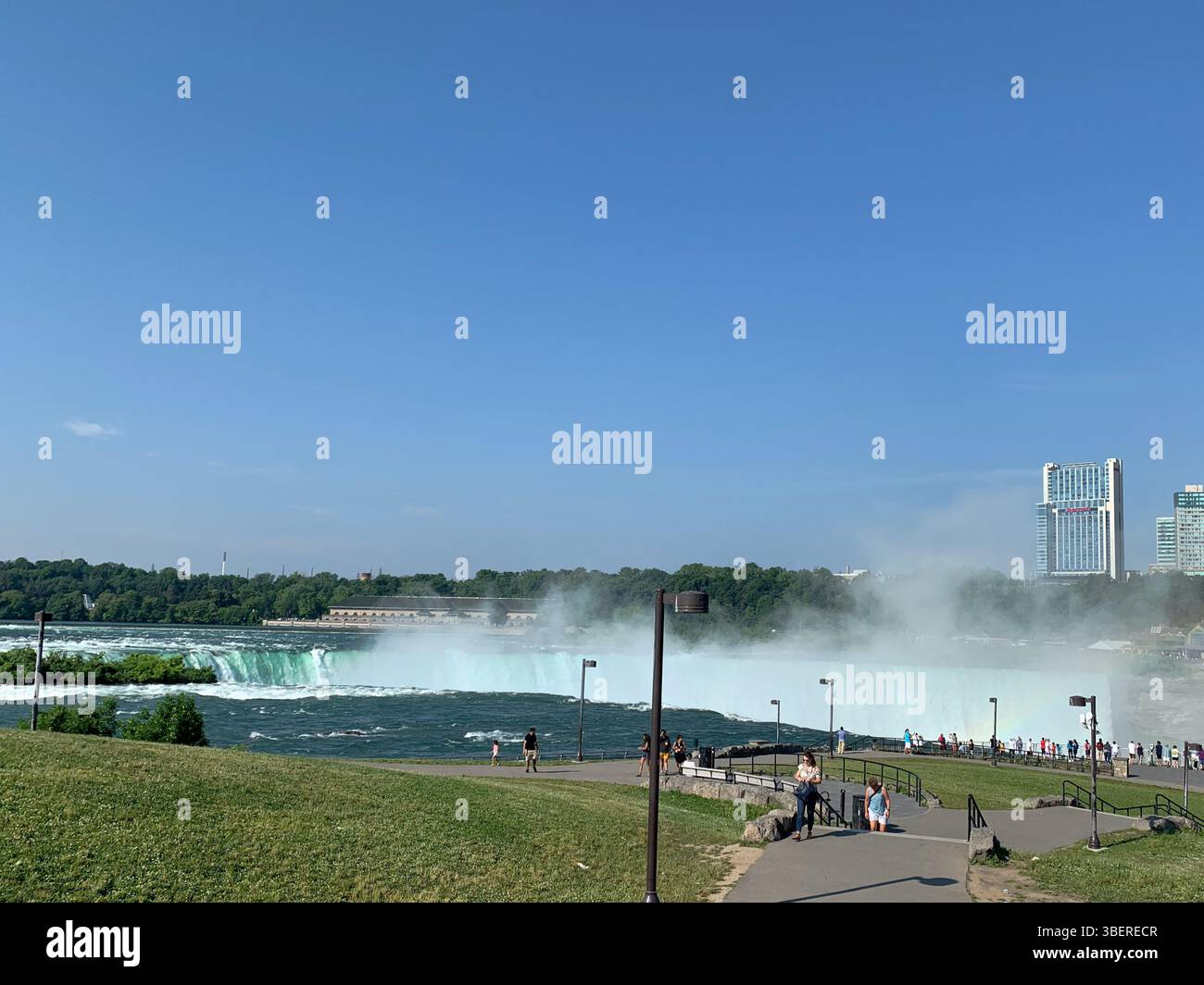 Touristen genießen die Aussicht auf die Niagarafälle und den Nebel, der von den Horseshoe Falls an einem klaren Sommertag von der US-Seite mit Kanadas Skyline aufsteigt. - Smartphone-aufgenommenes Stockfoto