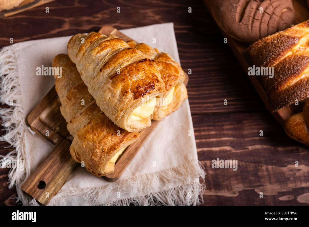 Eine verlockende Auswahl an frisch gebackenem Gebäck, mit cremigem Pudding gefülltem Blätterteig auf einem rustikalen Holzbrett. Stockfoto
