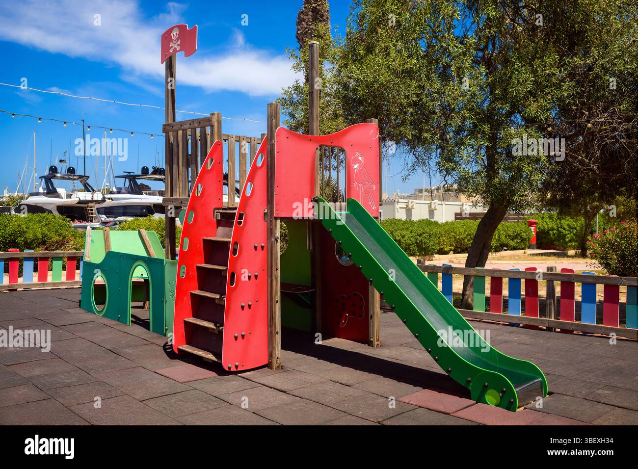 Kleine Spielanlage im Piratenstil mit Rutsche und Klettertreppen in einem sonnigen Park. Umgeben von Bäumen, farbenprächtigem Zaun und Yachten im Hintergrund. Stockfoto