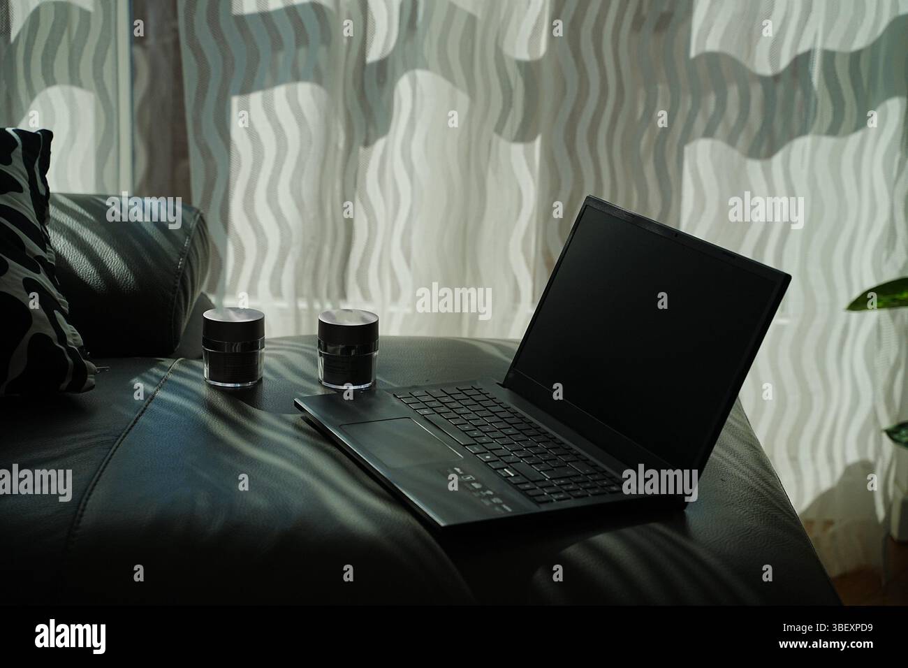 Laptop auf einer Ledercouch mit zwei Gläsern für Hautpflege-Creme. Gewellte Vorhänge werfen weiche Schatten auf die Einrichtung und schaffen so ein gemütliches und ordentliches Ambiente Stockfoto