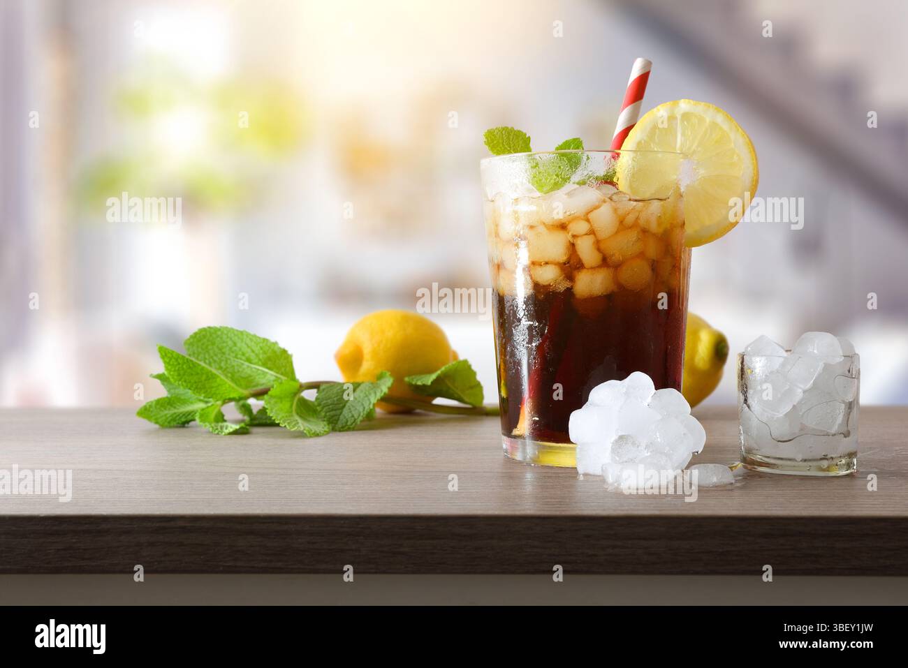 Erfrischendes Cola-Getränk mit Eis in einer Glasschale, dekoriert mit einem Strohhalm und einer Scheibe Obst auf einem Holztisch mit Obst, Eis und Minzblättern in lebendiger Atmosphäre Stockfoto