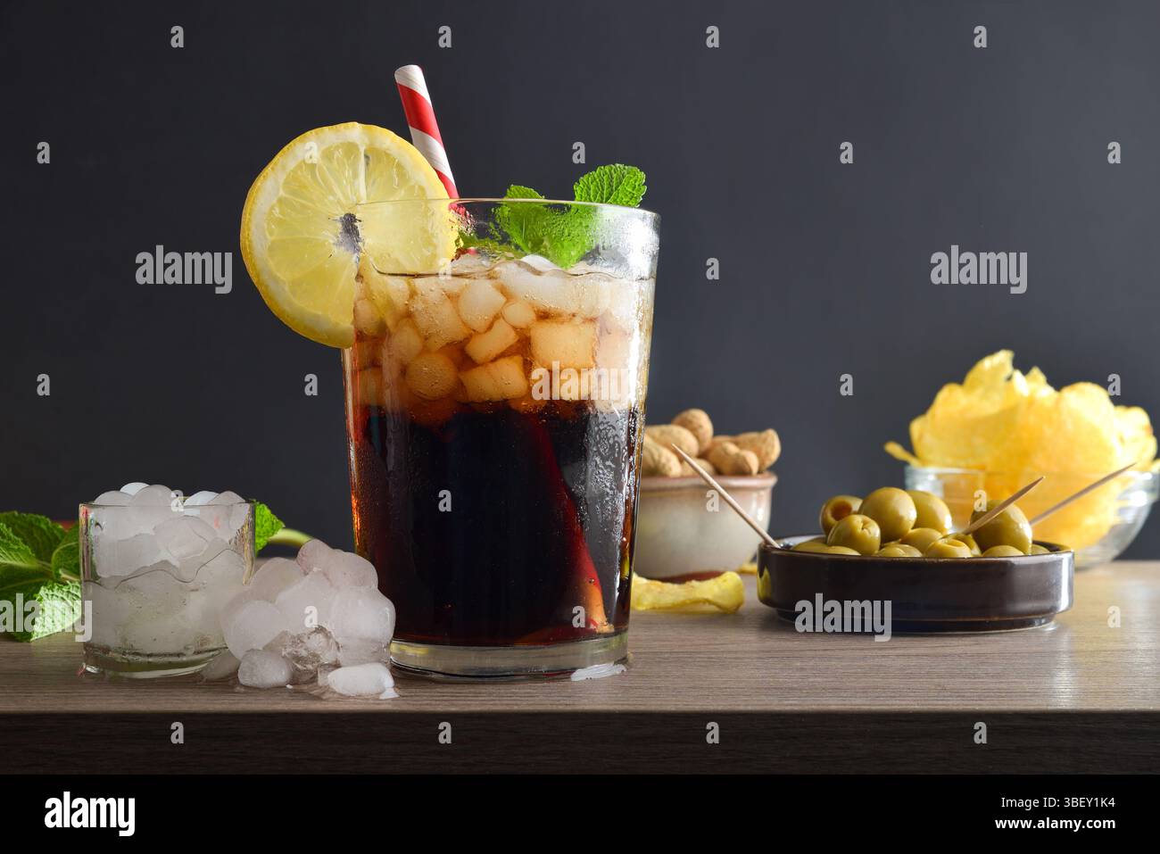 Erfrischendes Cola-Getränk auf einem Holztisch mit Snacks wie Erdnüssen und Oliven dekoriert mit Minzblättern und Zitronenscheiben isoliert mit schwarzem Hintergrund Stockfoto