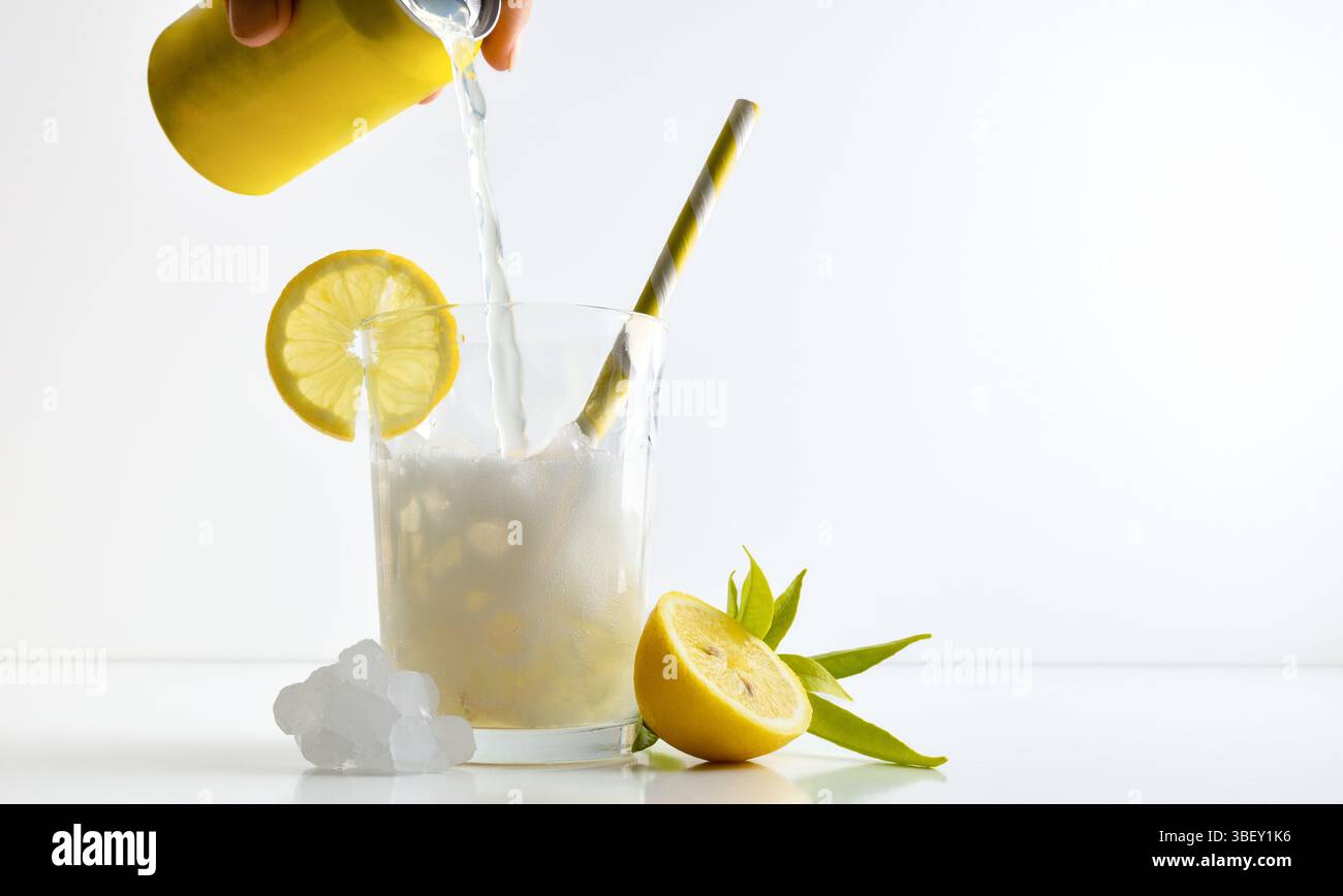 Erfrischendes Zitronengetränk mit Eis in einer Glasschale, dekoriert mit einem Strohhalm und einer Scheibe Obst auf einem weißen Tisch mit Obst, Eis und Blättern mit einem weißen IS Stockfoto