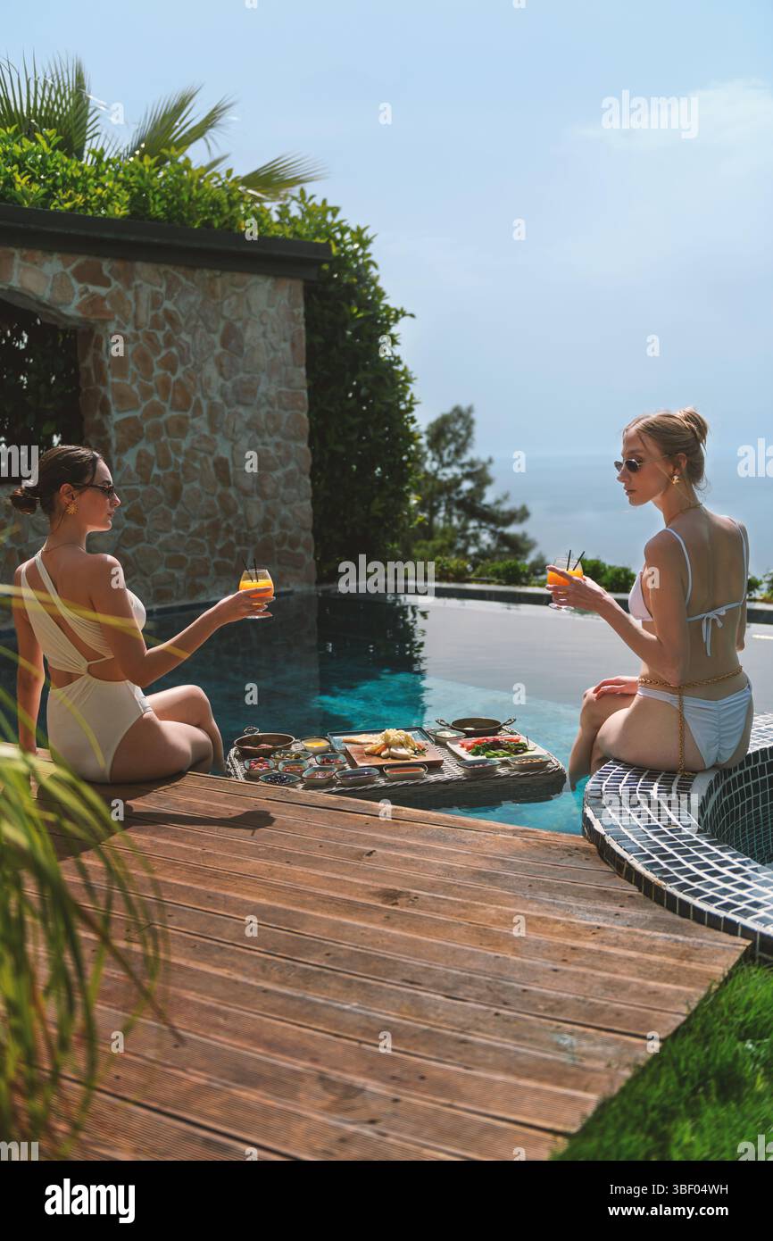 Eine Blondine und Brünette im Alter von 20 Jahren in Badeanzügen entspannen sich am privaten Pool, während sie ein schwimmendes Frühstück im Wasser genießen Stockfoto