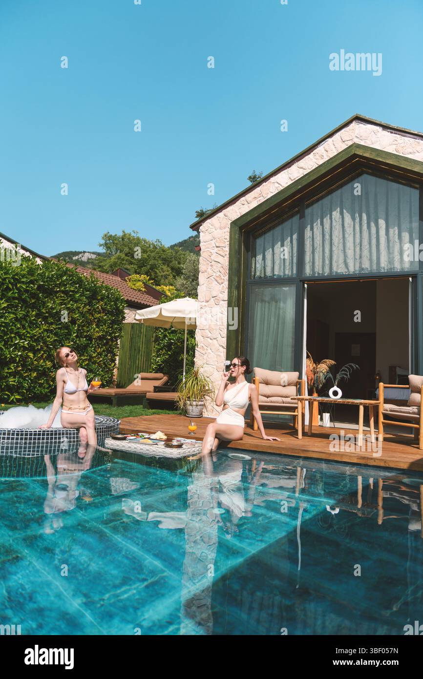 Eine Blondine und Brünette im Alter von 20 Jahren in Badeanzügen entspannen sich am privaten Pool, während sie ein schwimmendes Frühstück im Wasser genießen Stockfoto