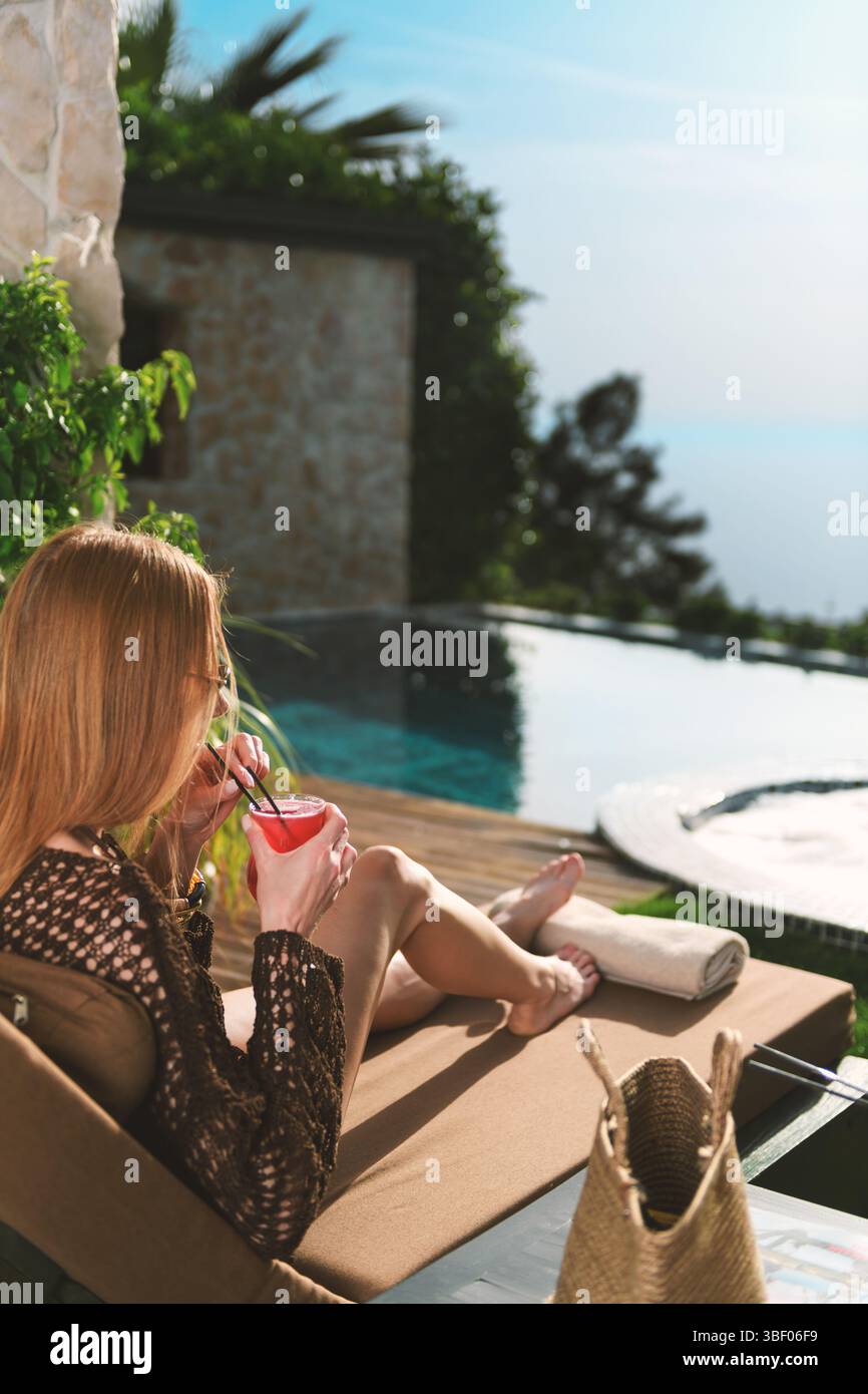 Eine blonde Frau aus den 20er Jahren in gestricktem Pareo und Sonnenbrille genießt ihren Cocktail, während sie die Aussicht bewundert, mit einem privaten Pool, der im Rahmen sichtbar ist. Stockfoto