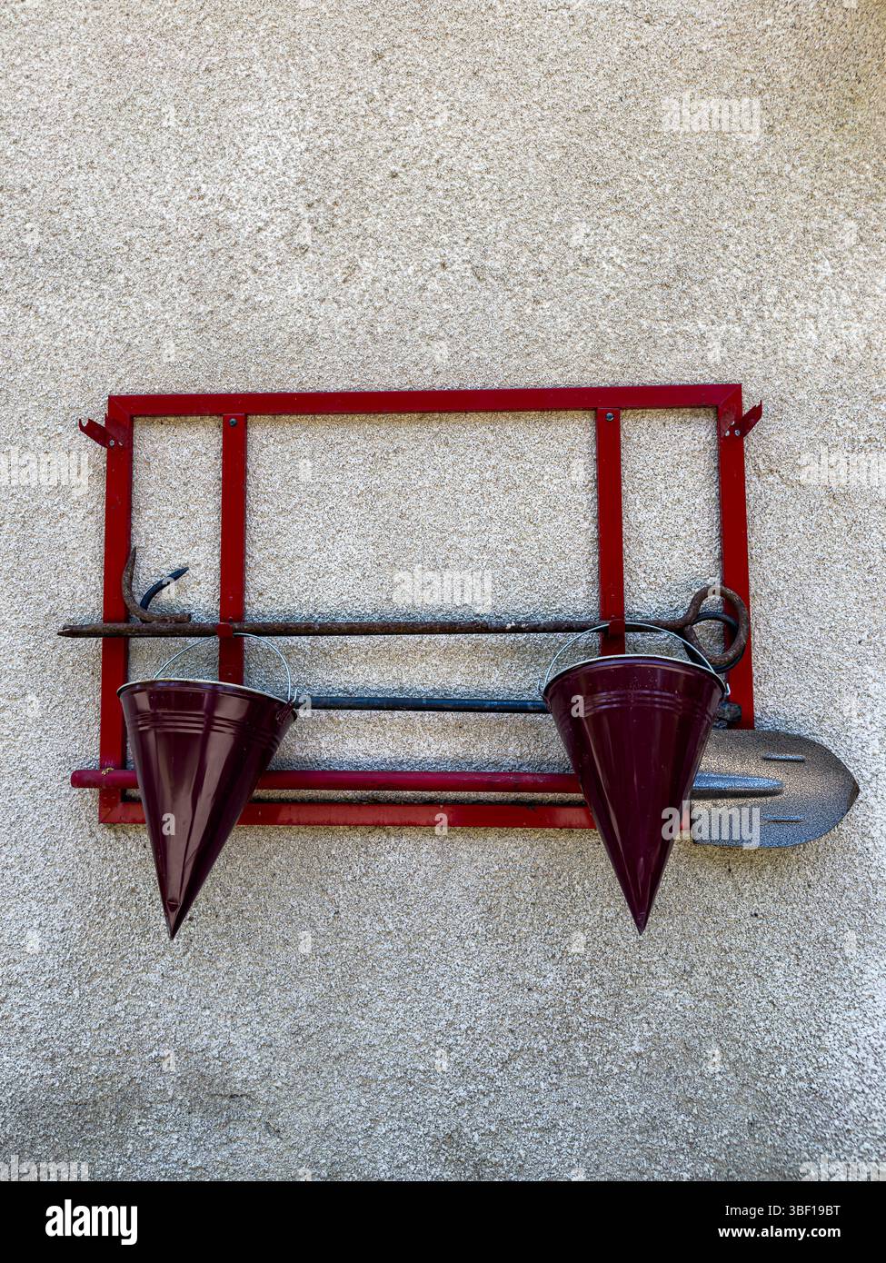 Löschausrüstung mit rotem Metallrahmen, Eimern, Schaufel und Haken, die an strukturierten Wänden hängen, Nahaufnahme, Tageslicht, Sicherheits- und Notfallkonzept. Stockfoto