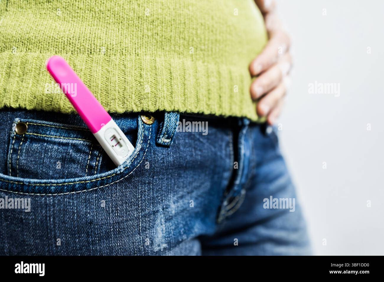 Nahaufnahme eines positiven Schwangerschaftstests, sichtbar in der Tasche der blauen Jeans, mit einer Hand sanft auf den Bauch gelegt, die eine frühe Schwangerschaft und Ameise symbolisiert Stockfoto