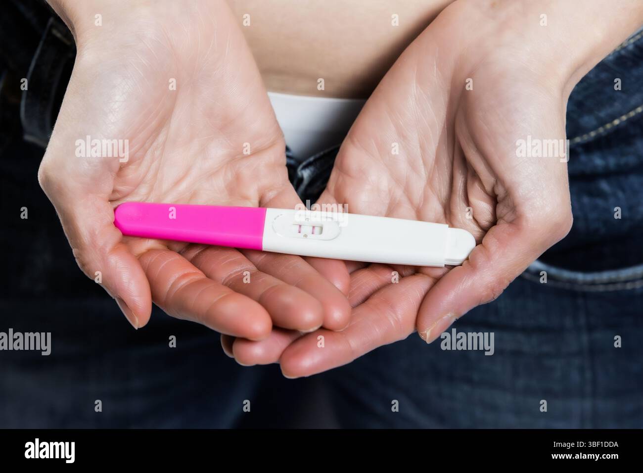 Detaillierte Ansicht der Hände einer Frau, die sanft einen positiven Schwangerschaftstest vor ihrem Bauch wiegen, was die emotionale Bedeutung eines frühen Schwangerschaftstests symbolisiert Stockfoto