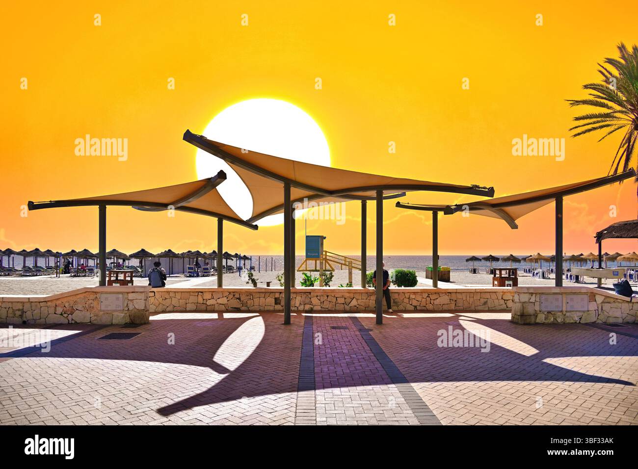 Der Strand von Malaga Fuengirola liegt vor orangefarbenem Himmel und der Sonne, die durch die Sonnenschirme platzt Stockfoto