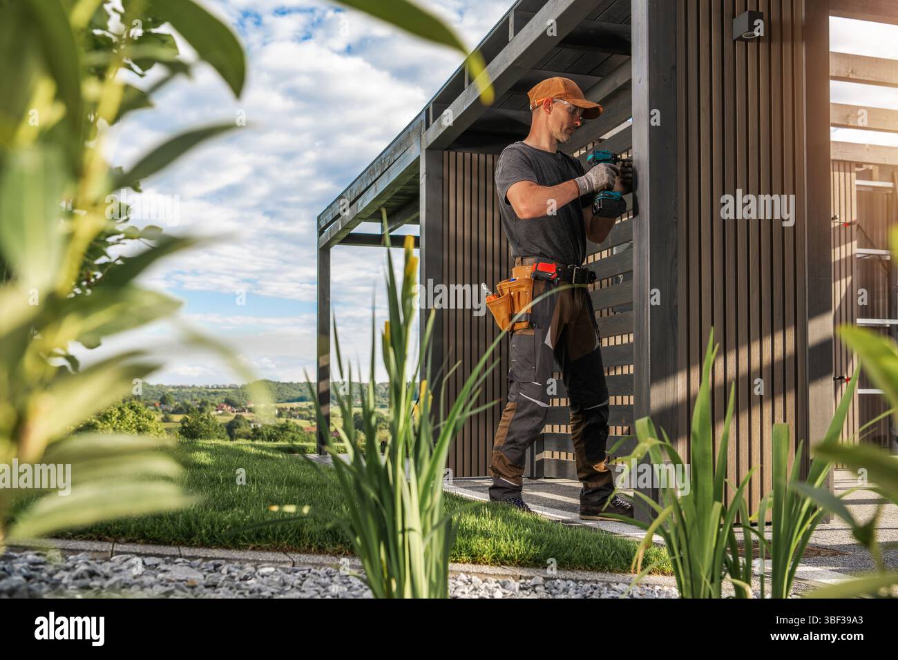 Ein Bauarbeiter installiert sorgfältig Elemente auf einer stilvollen Außenstruktur, umgeben von Grün und klarem Himmel. Der lebhafte Garten fügt eine neue Note hinzu Stockfoto