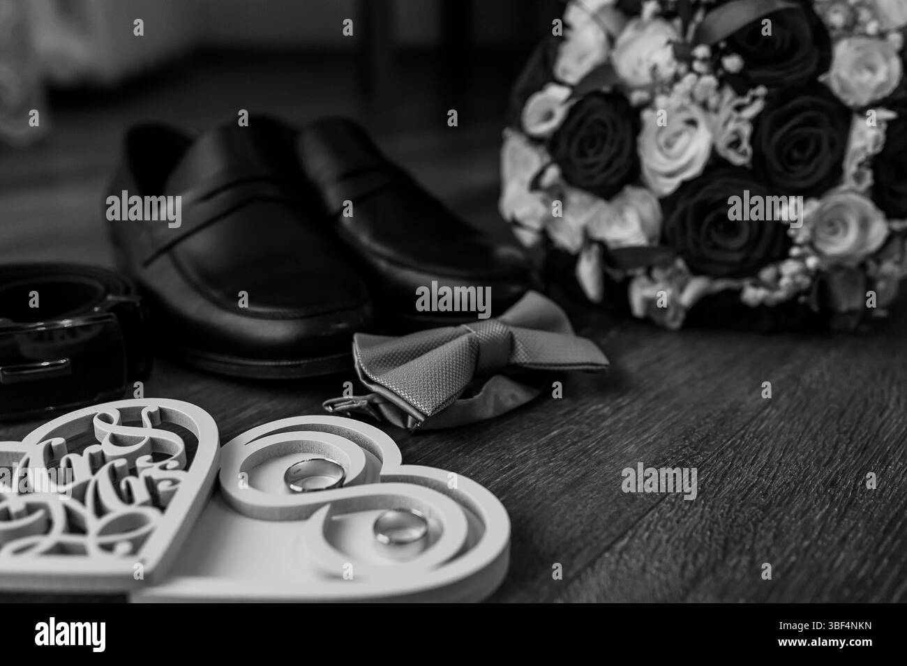 Elegantes Hochzeitsset mit polierten schwarzen Lederschuhen, einer grauen Schleife, einem atemberaubenden weißen und schwarzen Blumenstrauß und Hochzeitsringen, die auf einem warmen Stockfoto