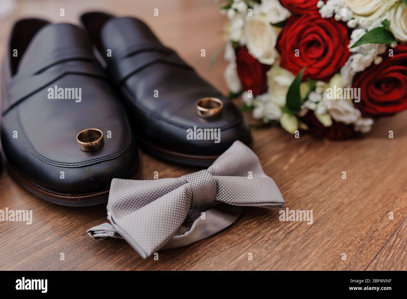 Eine wunderschön gestaltete Hochzeitsszene zeigt elegante schwarze Schuhe, goldene Ringe, die Engagement symbolisieren, eine strukturierte graue Schleife und einen atemberaubenden Blumenstrauß Stockfoto