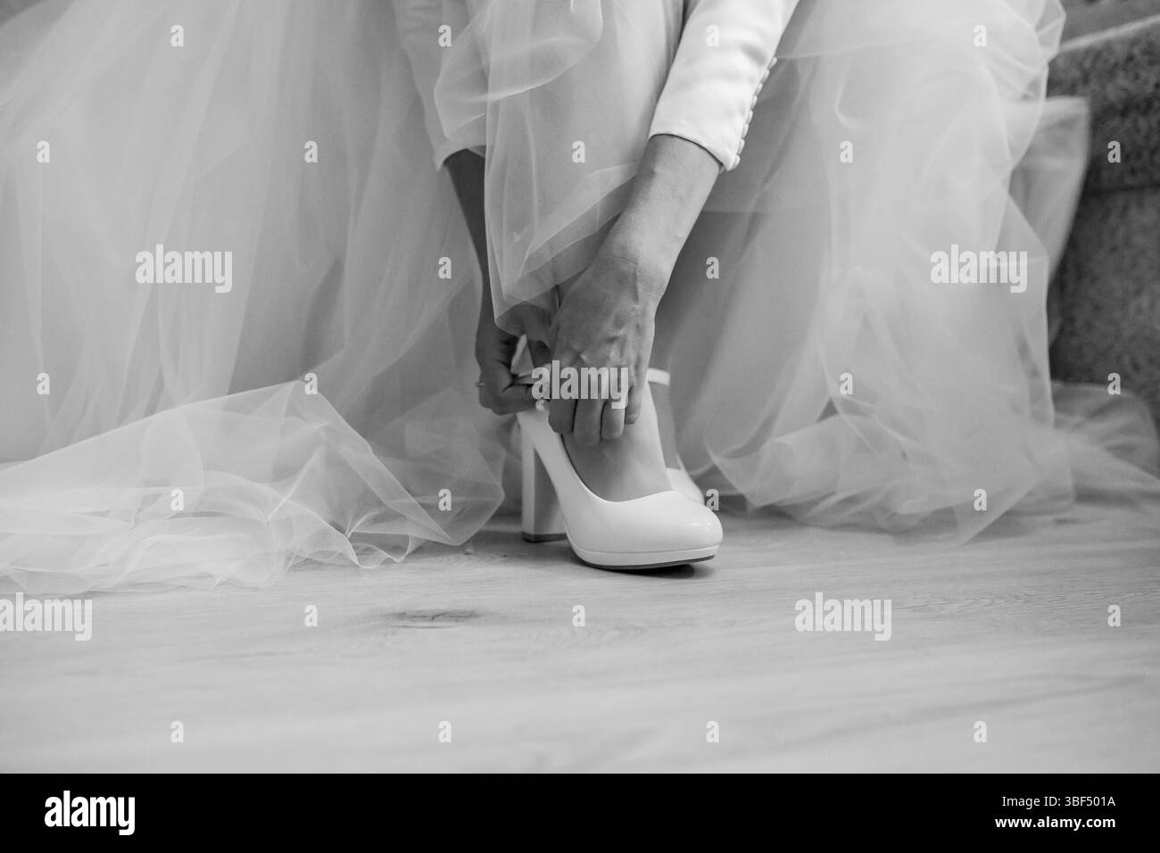 Ein monochromatisches Nahbild zeigt eine Braut, die sorgfältig weiße Hochzeitsschuhe anzieht, während sie ein fließendes Hochzeitskleid trägt, was ein Gefühl von Ser weckt Stockfoto
