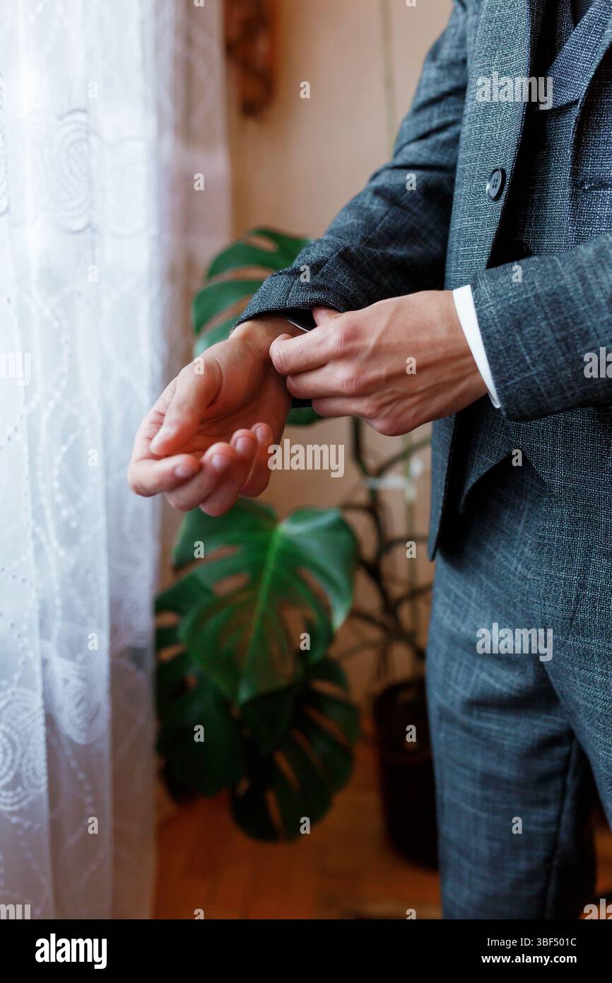 Ein Mann in einem maßgeschneiderten grauen Anzug mit Fischgrätmuster schnallt seine Manschettenknöpfe im Innenbereich akribisch, mit einer grünen Monsterapflanze als natürlicher Hintergrund Stockfoto