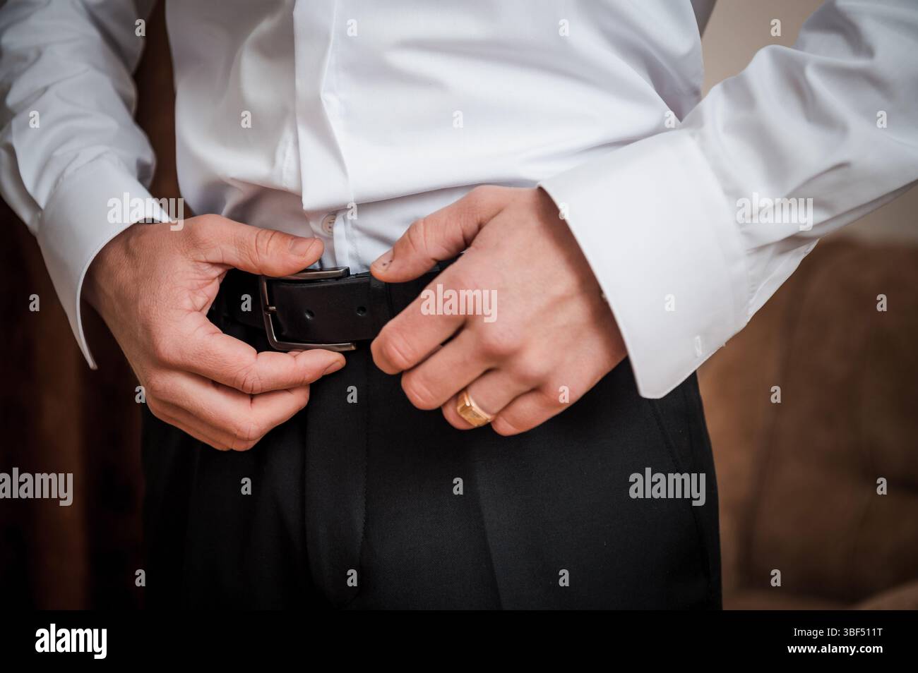 Eine Nahaufnahme zeigt einen Mann in einem weißen Hemd und einer schwarzen Hose, der einen Ledergürtel sichert, einen goldenen Ehering trägt, während der Hochzeitsvorbereitungen, Sugges Stockfoto