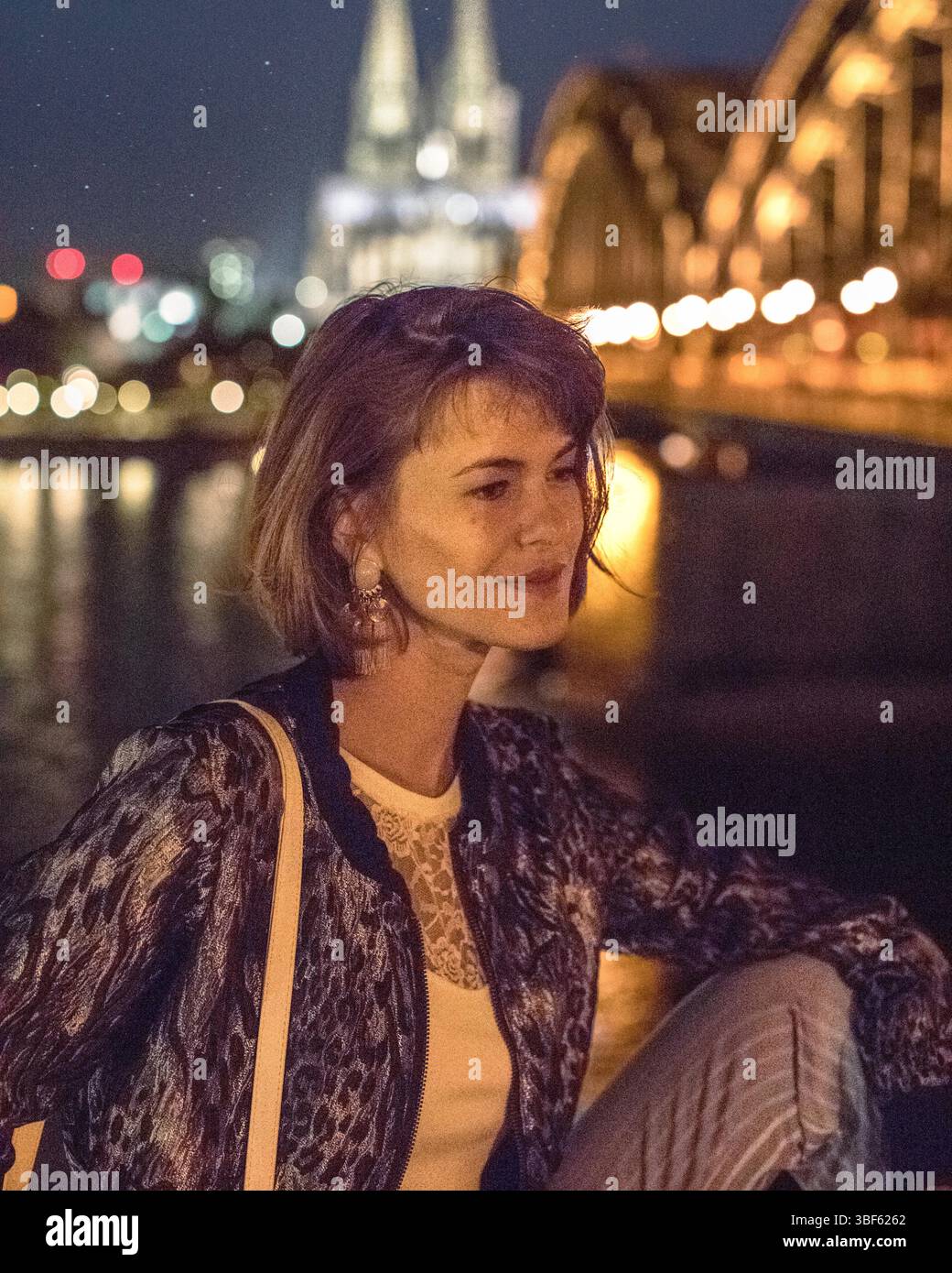 Portriat einer attraktiven jungen Frau vor der nächtlichen Skyline von Köln in Deutschland. Beleuchtete Brücke namens „die Hohenzollernbrücke“ Stockfoto