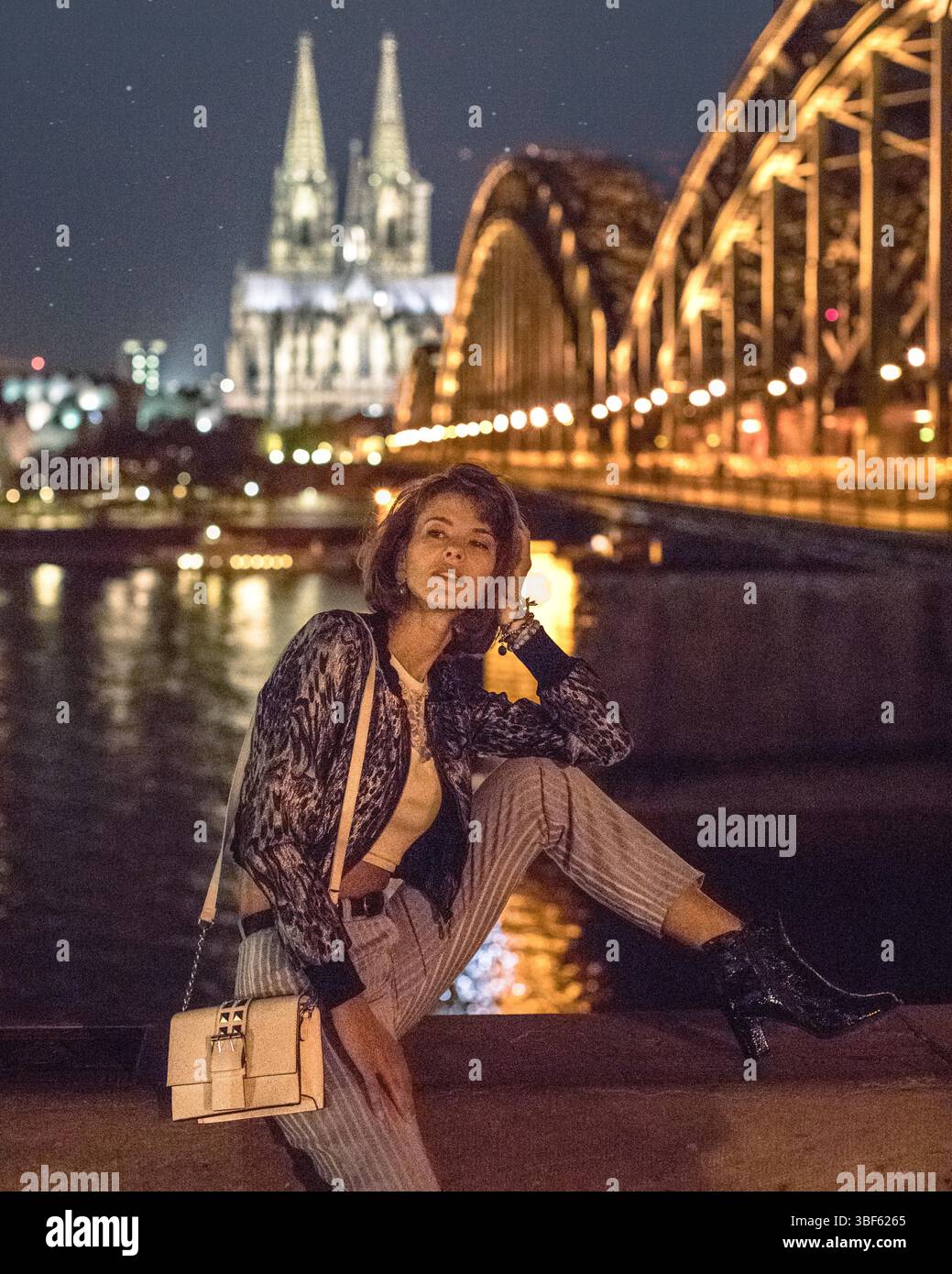 Attraktive junge Frau, die vor der nächtlichen Skyline von Köln in Deutschland sitzt. Beleuchtete Brücke, genannt „die Hohenzollernbrücke“ mit Kuppel. Stockfoto