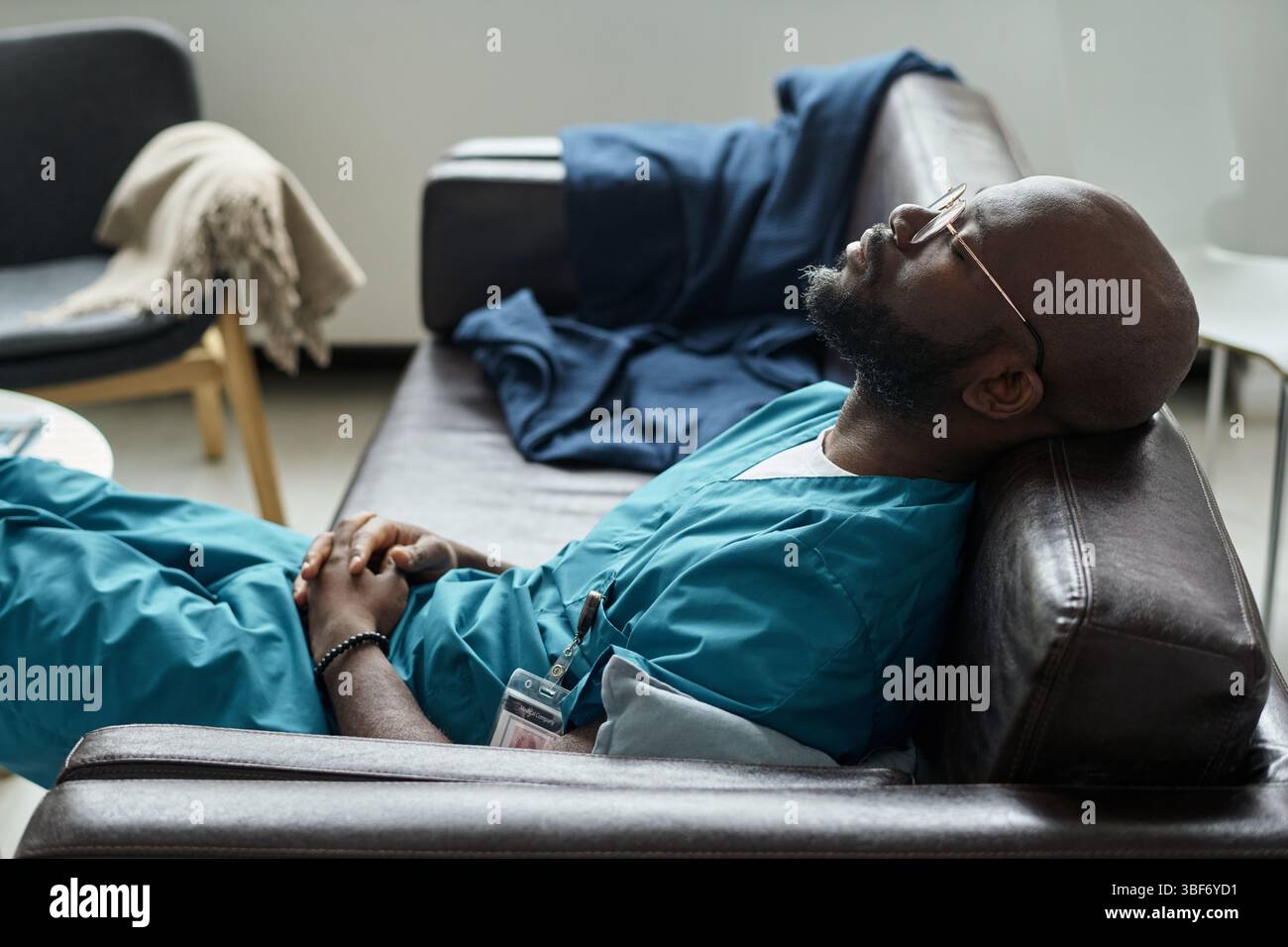 Die Krankenschwester mit Peelings ruht in einer Krankenhauslounge mit entspanntem Ausdruck, auf einem Sofa mit einer Decke und anderen persönlichen Gegenständen in der Nähe Stockfoto