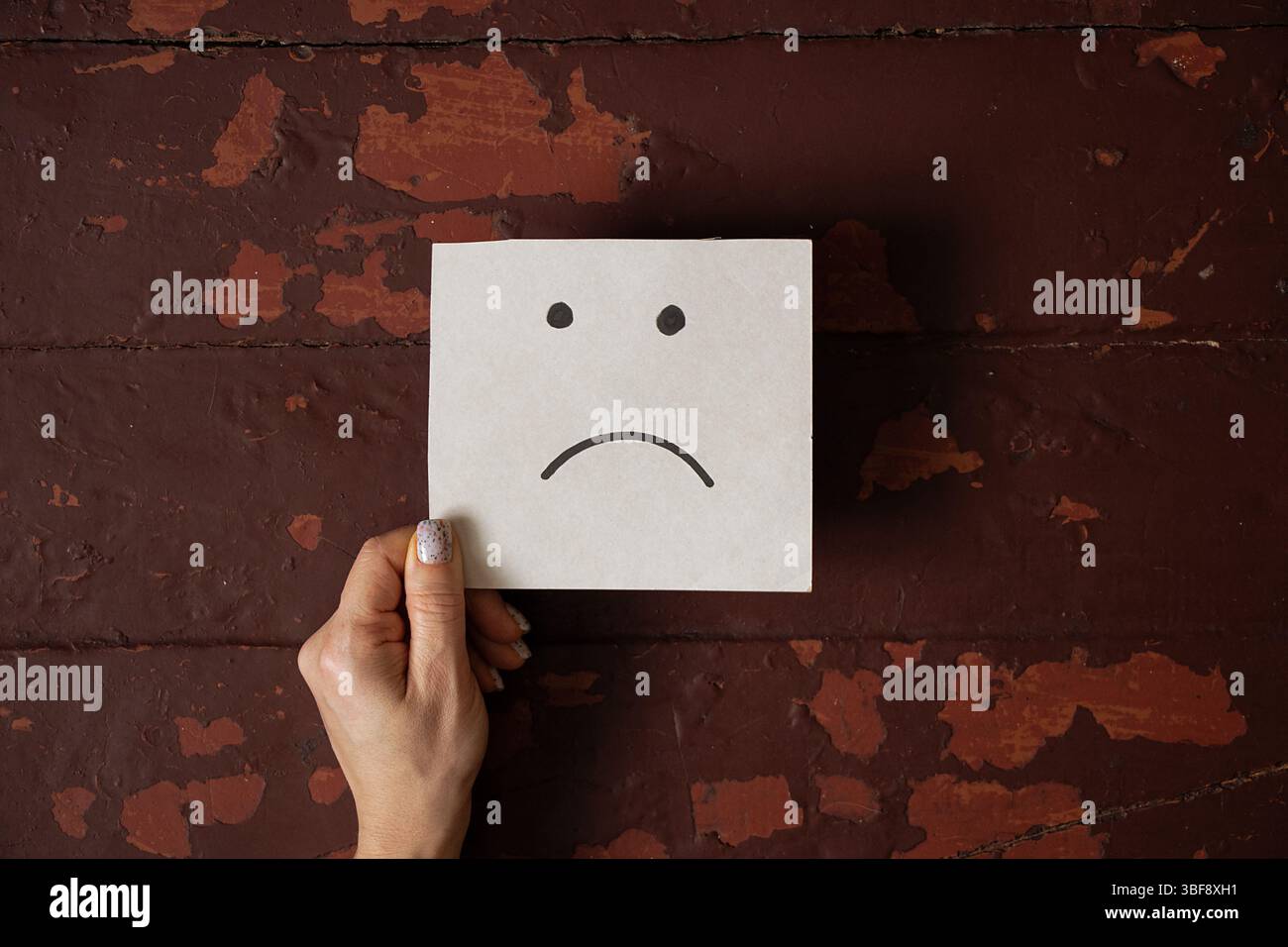 Weibliche Hand, die eine Karte mit einem traurigen Gesicht auf dem Hintergrund eines alten braunen Holztisches hält, Emotion von Traurigkeit und Unzufriedenheit Stockfoto