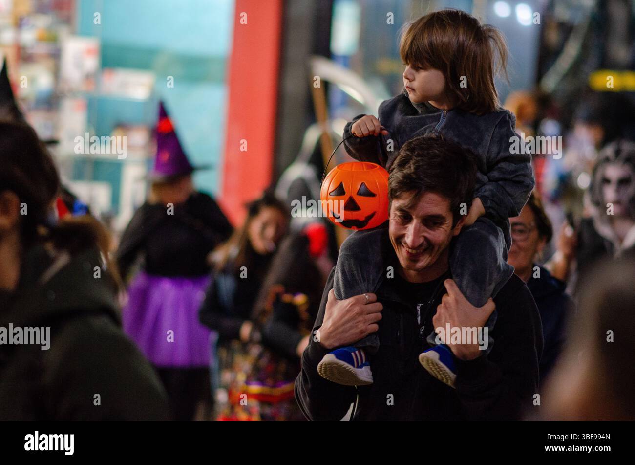 Der fröhliche Vater trägt sein Kind während eines lebhaften spanischen Halloween-Straßenkarnevals auf den Schultern, beide genießen festliche Kostüme und helle Kürbislant Stockfoto