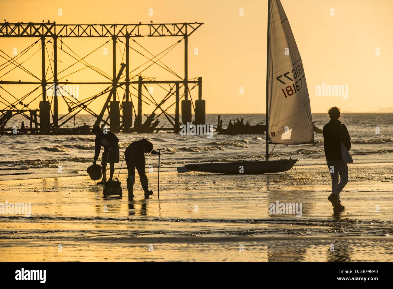 Fischer mit Eimern, die bei Ebbe in den Sand graben, um Würmer als Köder zu verwenden. Ein kleines Segelboot und die verfallene Struktur des West Pier im Hintergrund. Brighton Beach. England. Silhouetten. Brighton, England, Großbritannien. Stockfoto