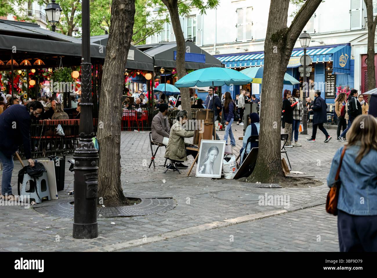 Pulsierende Straßenszene in einer europäischen Stadt, wahrscheinlich Paris, mit Künstlern, die unter bunten Regenschirmen Porträts zeichnen, und Menschen, die ein Café im Freien genießen Stockfoto