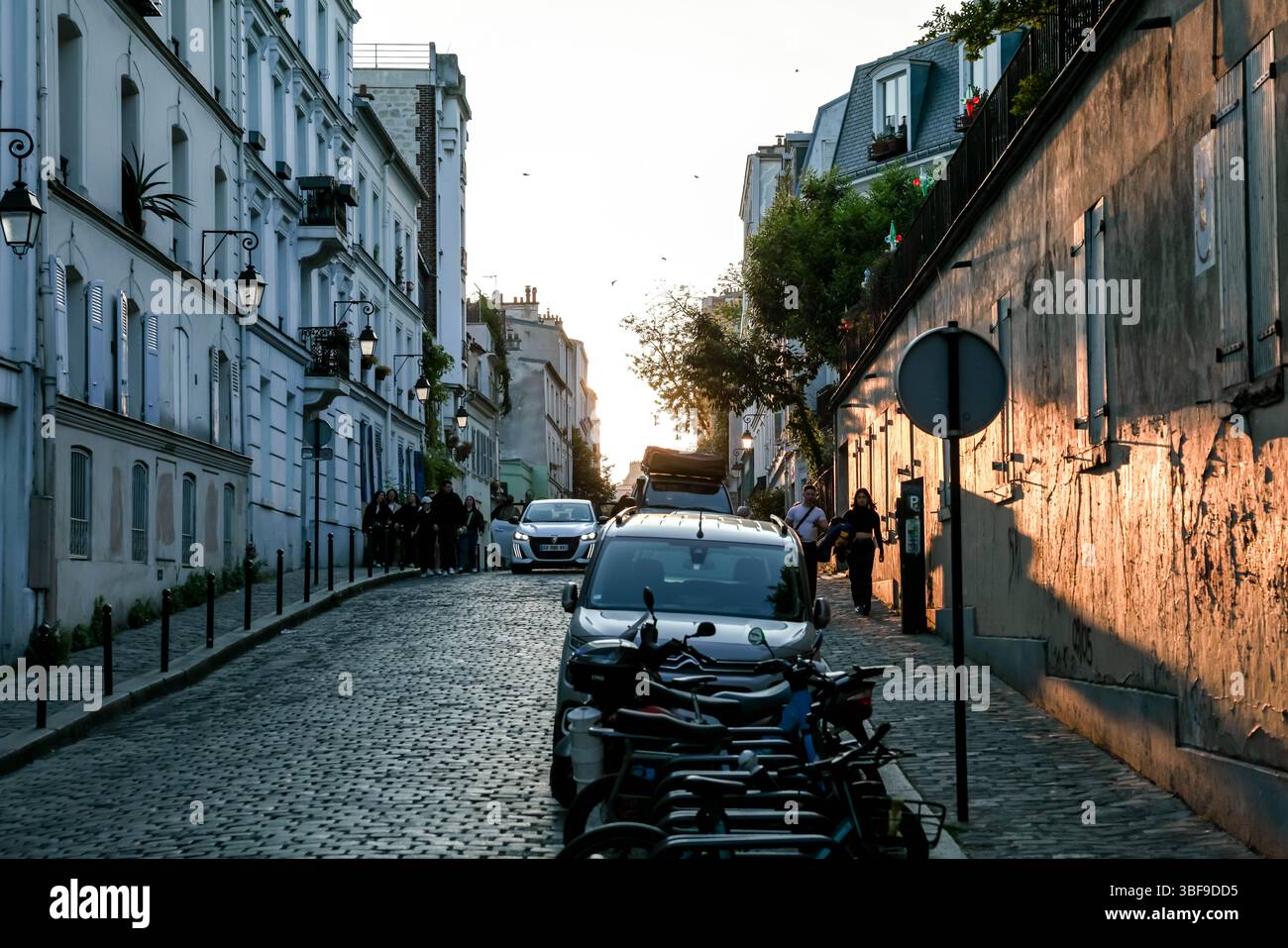 Dieses Bild zeigt eine bezaubernde Pariser Kopfsteinpflasterstraße bei Sonnenuntergang, mit geparkten Rollern, die den Weg säumen und warmem Licht, das lange Schatten wirft Stockfoto