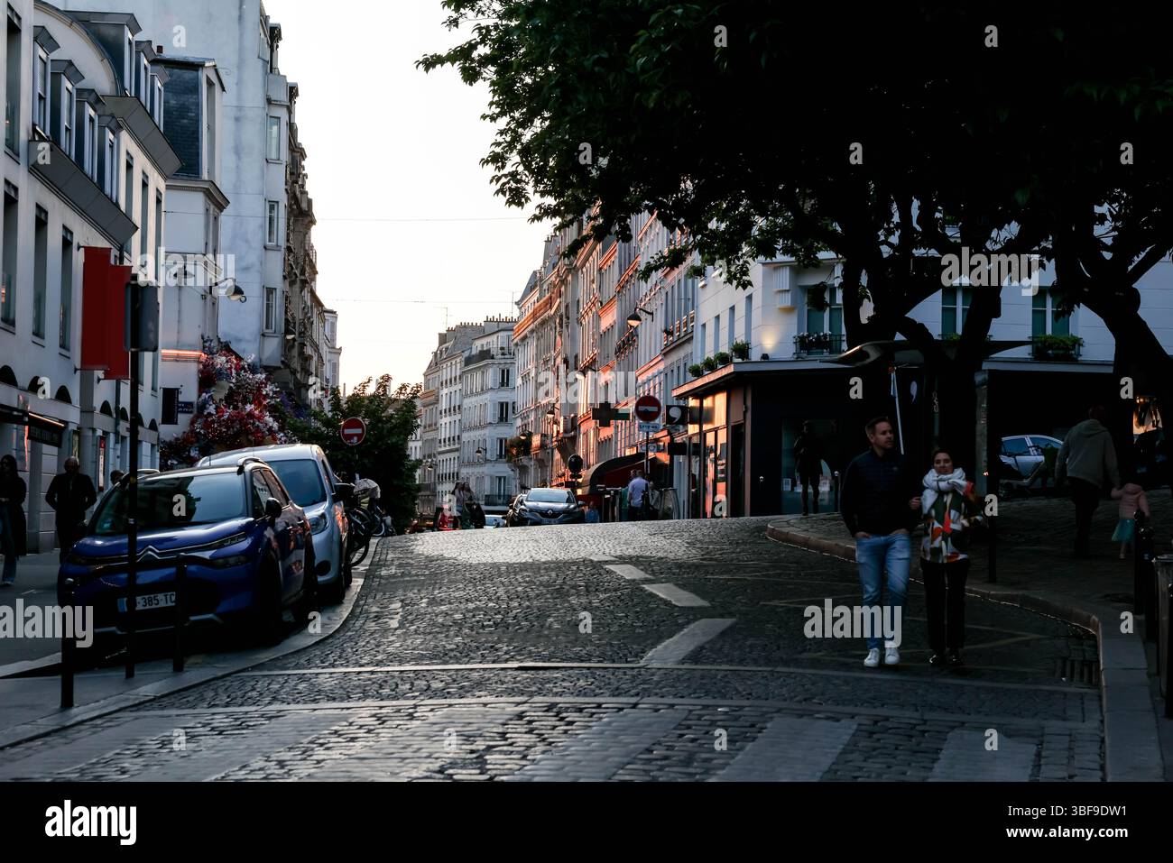 Ein Paar genießt einen gemütlichen Spaziergang entlang der kopfsteingepflasterten Rue des Abbesses im Montmartre-Viertel von Paris, während die Dämmerung sich mit historischem Gebäude einsetzt Stockfoto