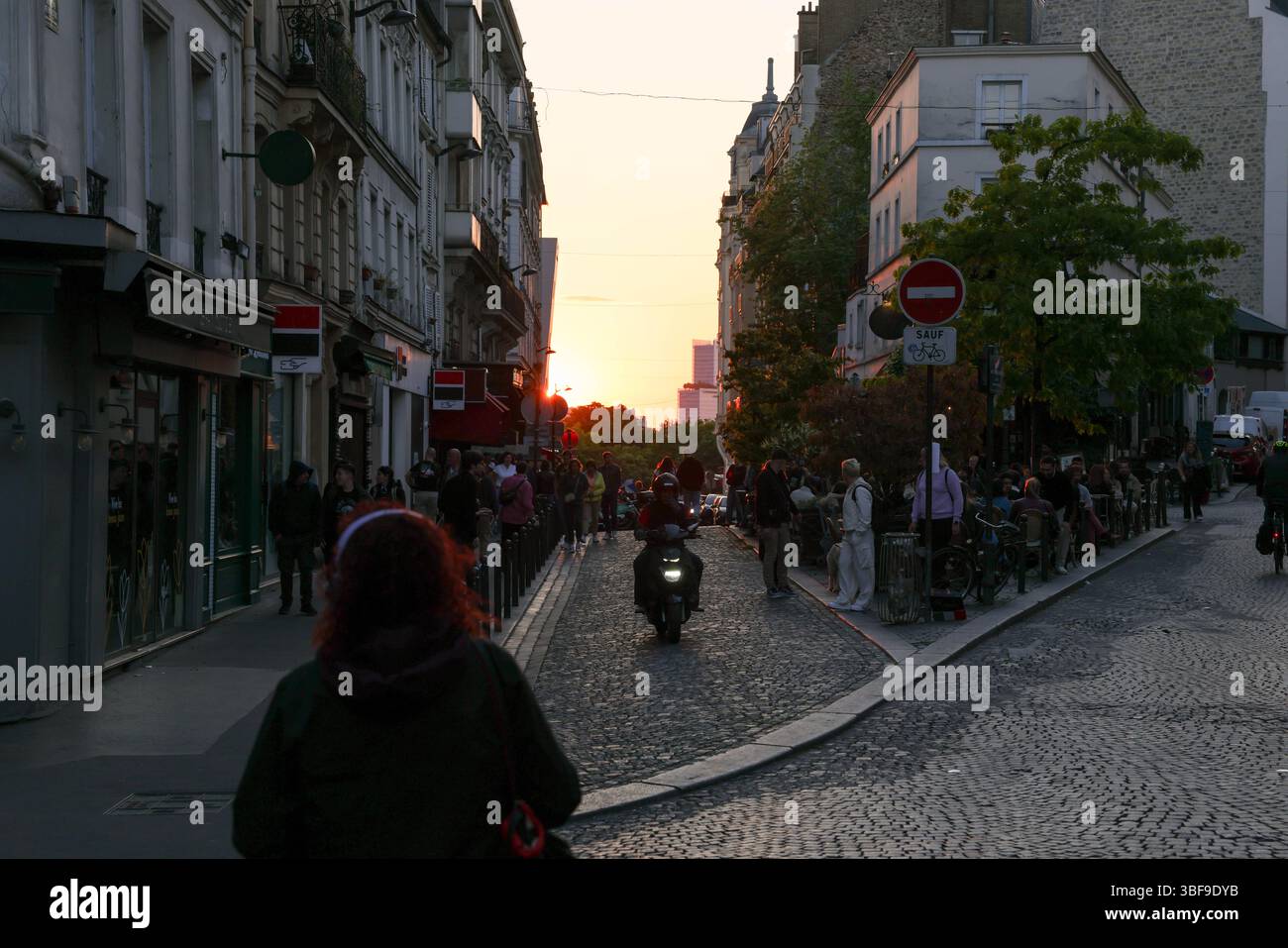 Blicken Sie auf eine kopfsteingepflasterte Pariser Straße direkt in die untergehende Sonne und schaffen Sie Silhouetten von Gebäuden und Menschen, die das warme Leuchten einfangen Stockfoto