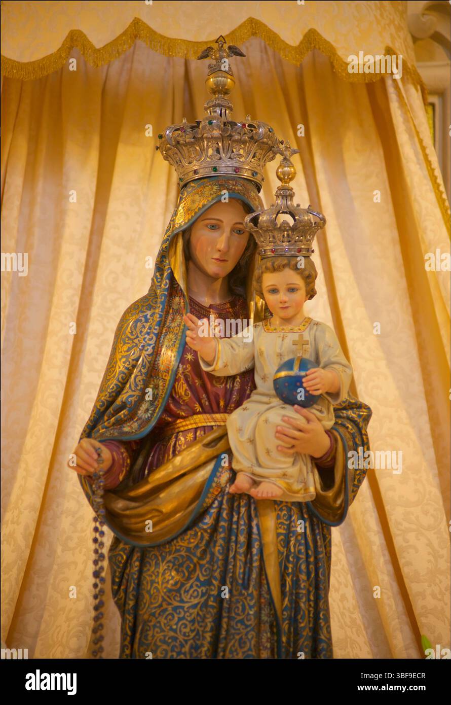 Statue der Jungfrau Maria, die das Jesuskind hält. Beide Figuren sind mit kunstvollen Kronen verziert. Die Jungfrau Maria trägt ein blau-goldenes Gewand, während das Jesuskind weiß gekleidet ist und eine Kugel hält. Die Statue scheint aus bemaltem Gips oder ähnlichem Material zu bestehen und ist vor einem Hintergrund aus drapiertem Stoff gestellt. Die Kronen auf beiden Figuren sind metallisch und mit Steinen verziert. Nardo, Apulien, Italien Stockfoto