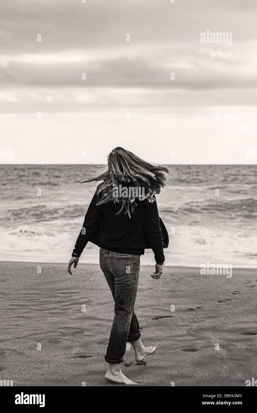 Junge Frau, die barfuß am Strand entlang läuft und zarte Spuren im weichen Sand hinterlässt, während die Wellen des Meeres sanft am Ufer liegen. Stockfoto