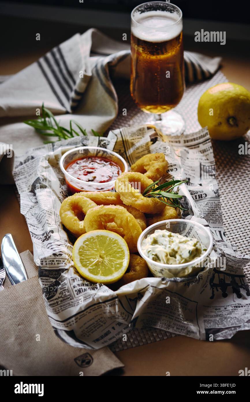 Nahaufnahme knuspriger Calamari-Ringe, serviert mit eintauchenden Soßen, Zitronenscheibe und Kräutern auf Zeitungspapier, mit einem Glas Bier im Hintergrund Stockfoto