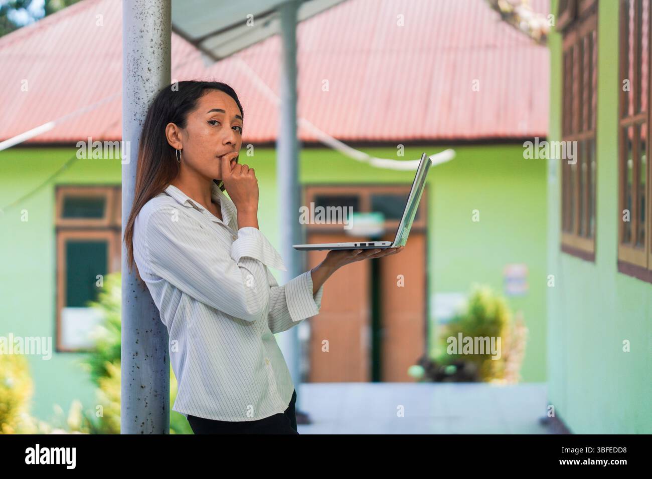 Eine nachdenkliche junge Frau, die einen offenen Laptop hält, sich auf eine Säule stützt, während sie in die Kamera blickt. Stockfoto