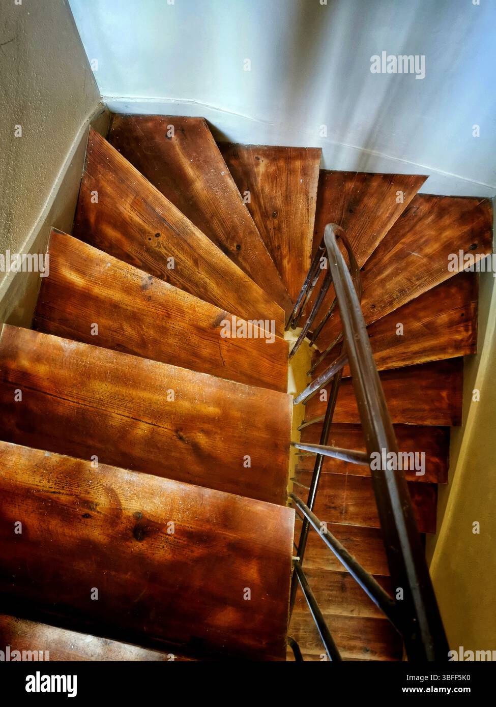 Eine hölzerne Wendeltreppe führt in eine helle untere Ebene hinab und zeigt satte Kornmuster und ein elegantes Metallgeländer. - Smartphone-aufgenommenes Stockfoto