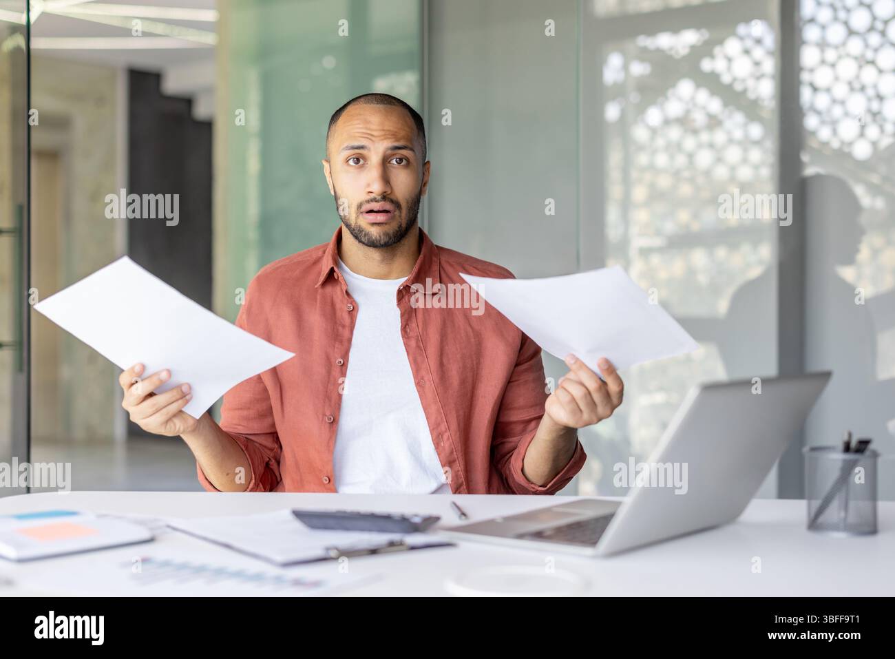 Ein überraschter Mann hält Papierkram, während er an seinem Schreibtisch arbeitet, und hat möglicherweise finanziellen Stress. Stockfoto