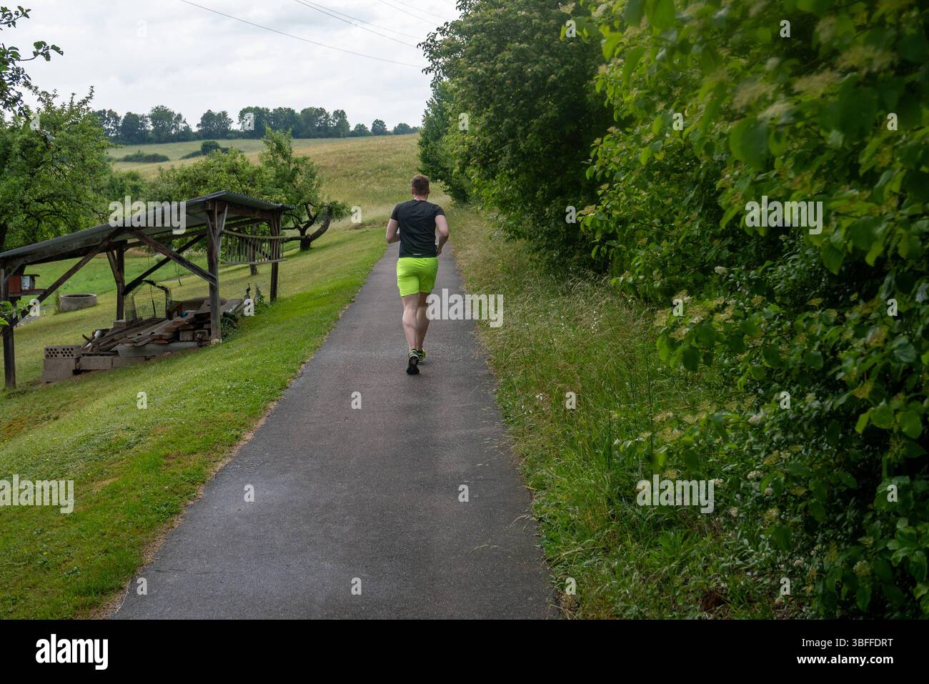 Jogger auf ländlichem Weg im Grünen 01.06.25, Selters: Symbolfoto, Illustrationsbild, Symbolbild, Illustrationsfoto, Alltagsszene ein Mann joggt auf einem schmalen asphaltierten Weg durch eine grüne, leicht hügelige Landschaft. Links des Weges stehen einfache Holzunterstände und Wiesen mit Obstbäumen, rechts dichter Busch- und Baumbewuchs. Der Himmel ist bewölkt, das Licht weich. Der Jogger trägt neongelbe Shorts und ein dunkles Sportshirt. Selters *** Jogger auf einem ländlichen Weg auf dem Land 01 06 25, Selters Symbolfoto, Illustrationsfoto, Symbolfoto, Illustrationsfoto, Alltagsszene Stockfoto