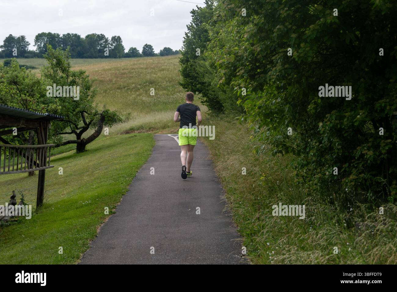 Jogger auf ländlichem Weg im Grünen 01.06.25, Selters: Symbolfoto, Illustrationsbild, Symbolbild, Illustrationsfoto, Alltagsszene ein Mann joggt auf einem schmalen asphaltierten Weg durch eine grüne, leicht hügelige Landschaft. Links des Weges stehen einfache Holzunterstände und Wiesen mit Obstbäumen, rechts dichter Busch- und Baumbewuchs. Der Himmel ist bewölkt, das Licht weich. Der Jogger trägt neongelbe Shorts und ein dunkles Sportshirt. Selters *** Jogger auf einem ländlichen Weg auf dem Land 01 06 25, Selters Symbolfoto, Illustrationsfoto, Symbolfoto, Illustrationsfoto, Alltagsszene Stockfoto