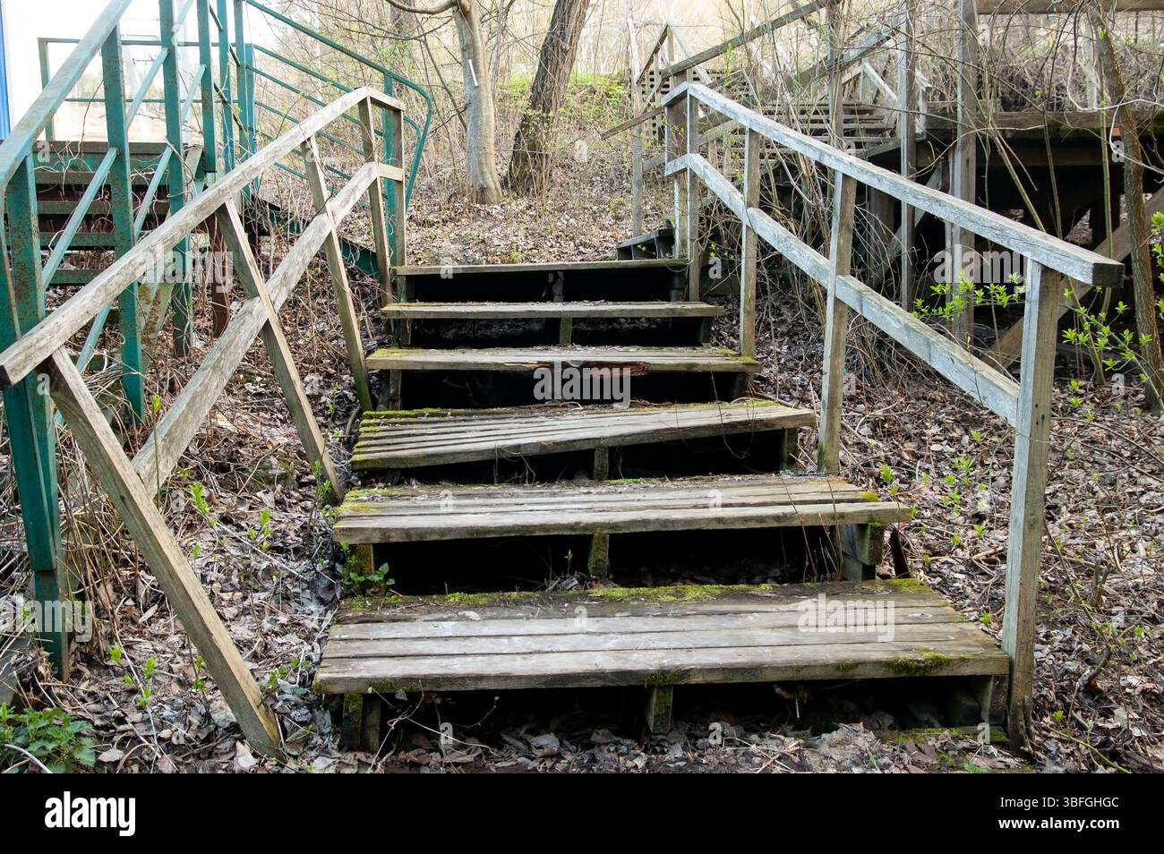 Eine verfallene Holztreppe von unten gesehen, mit Flecken von grünem Moos auf einigen Stufen. Daneben befindet sich eine rostige Metalltreppe mit Spuren von verblasstem grünem p Stockfoto