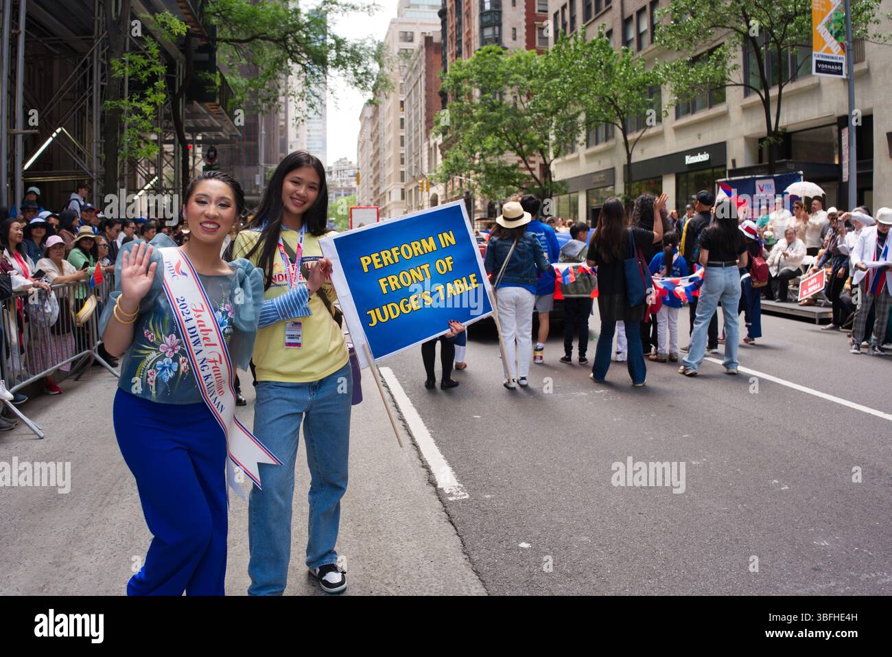 Zwei junge Frauen, eine mit Schärpe, stehen während einer Parade in einer Stadtstraße und halten ein Schild mit der Aufschrift "vor dem Tisch des Richters auftreten". Viele davon Stockfoto