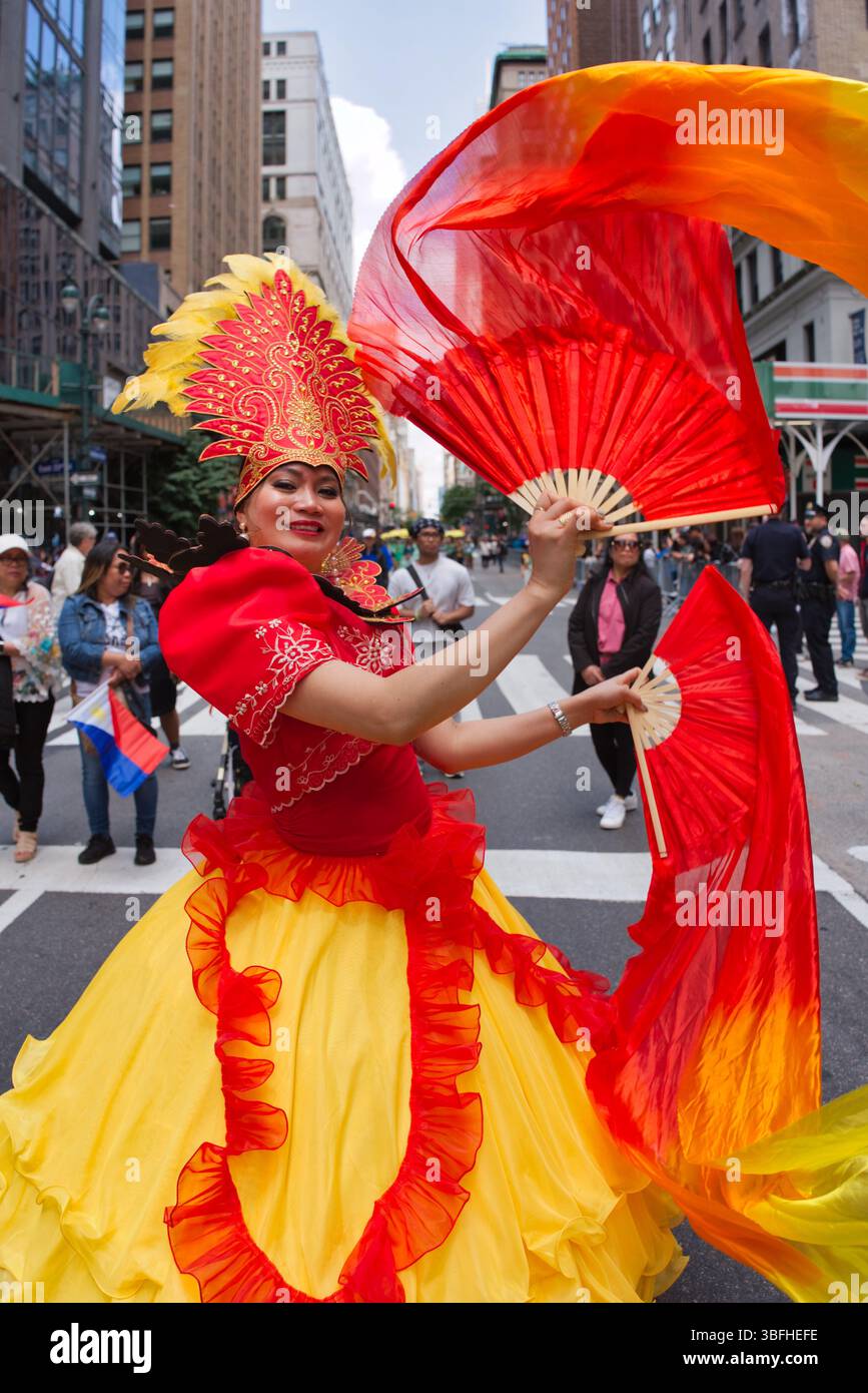 Eine Frau in einem leuchtend gelb-roten Kleid, verziert mit einem gefiederten Kopfschmuck, tritt mit großen roten und orangen Fans während einer Parade mit Stadtbügel auf Stockfoto