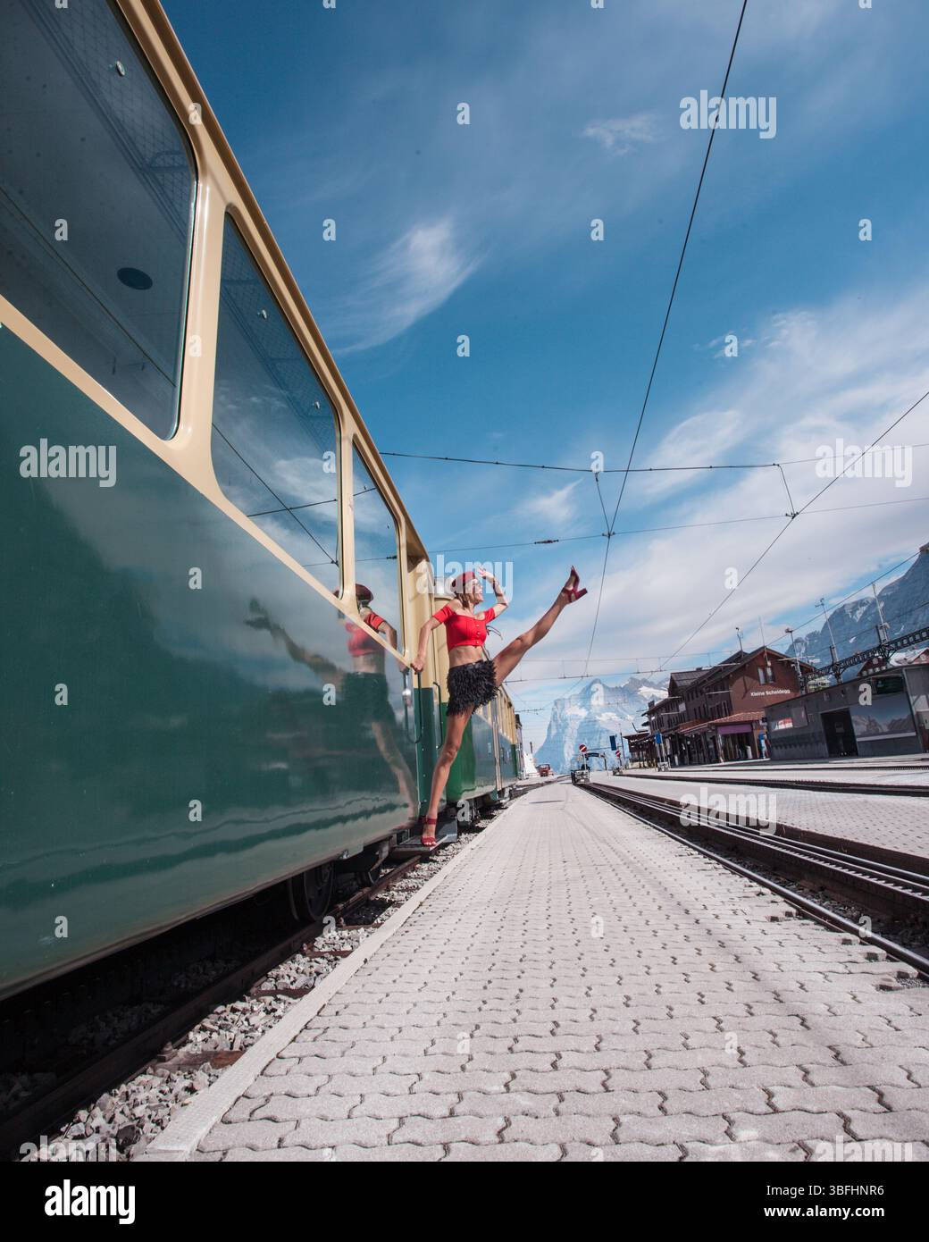 Modemodell, das mit einem Bein nach oben aus dem alten grünen Retro-Zug kommt, wie ein Turner oder Balletttänzer. Im Hintergrund Bergpass kleine Scheidegg Stockfoto