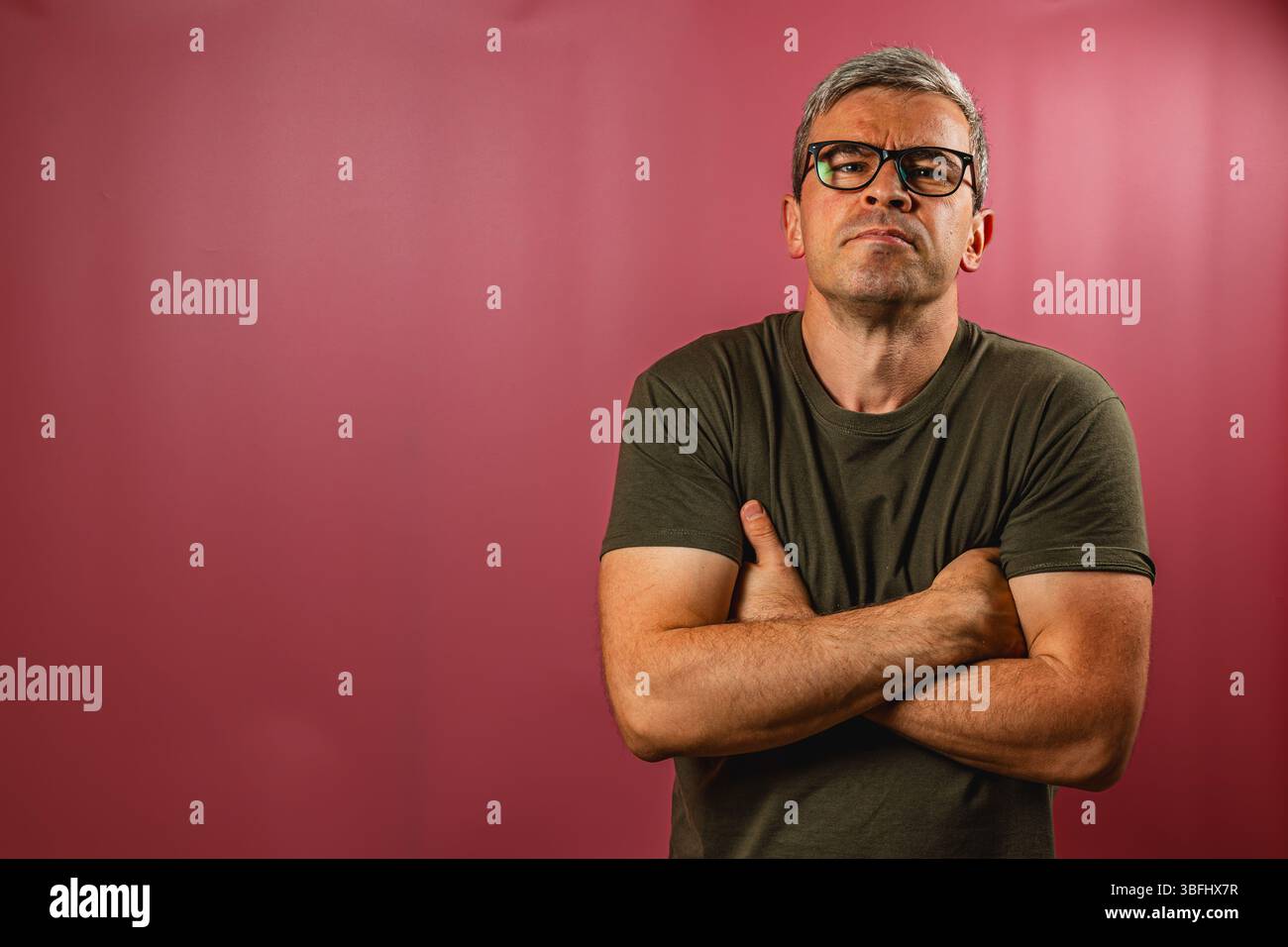 Ein Mann steht selbstbewusst mit gekreuzten Armen, trägt eine Brille und ein lässiges Hemd. Er blickt nachdenklich und zeigt einen kontemplativen Ausdruck gegen A Stockfoto
