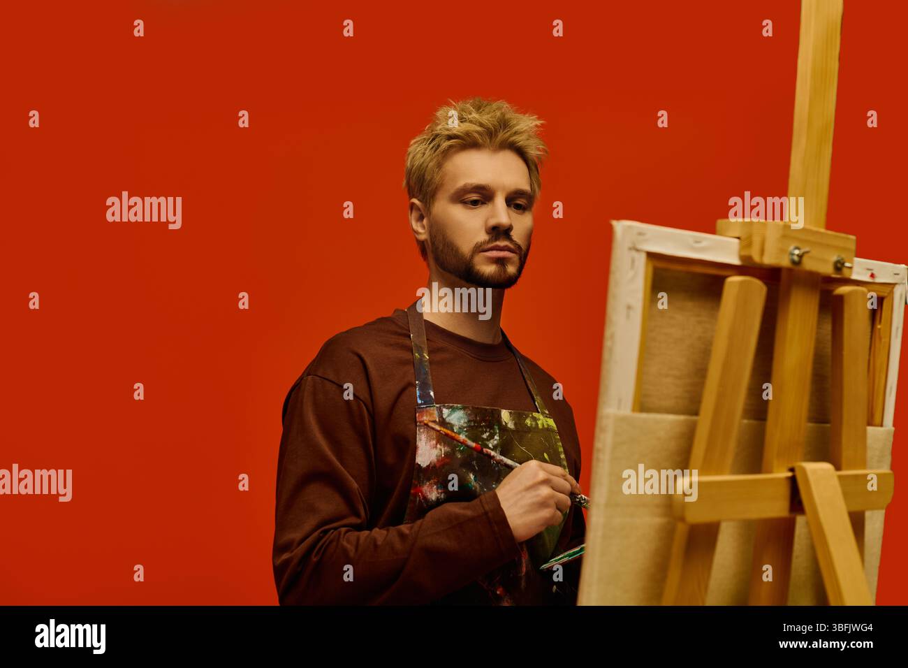Junger Mann, der sich mit der Malerei beschäftigt, zeigt künstlerischen Geist in einer markanten roten Atelierumgebung. Stockfoto