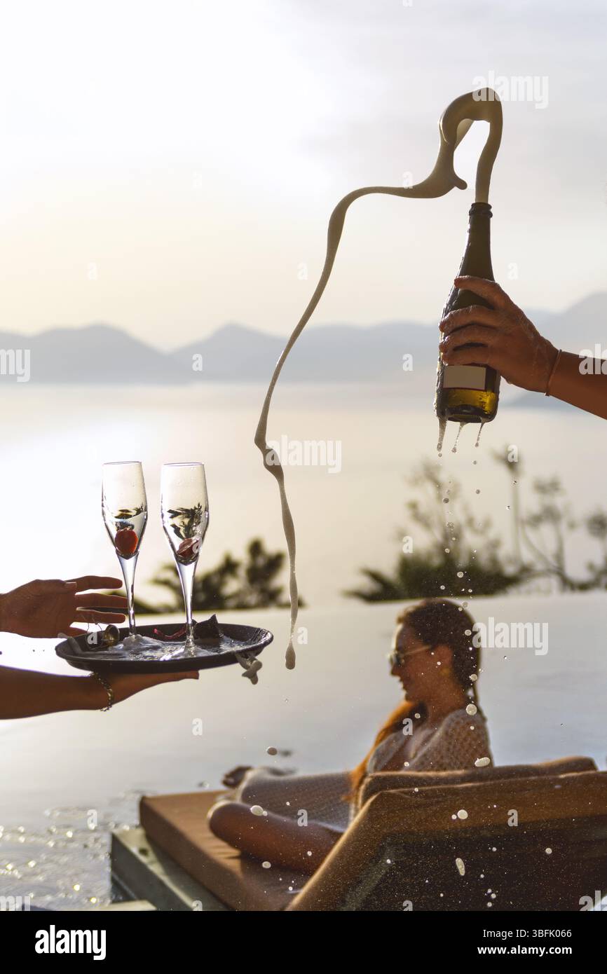 Eine Brünette aus den 20er Jahren loungiert sich bei Sonnenuntergang auf einer Sonnenliege am Pool, während der Champagner geplatzt wird und das Brausespray der Flasche scharf einfängt. Stockfoto