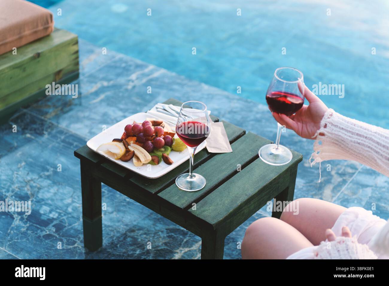 Eine intime Nahaufnahme von zwei Rotweingläsern und einer kunstvoll gestalteten Obstplatte am Pool. Stockfoto