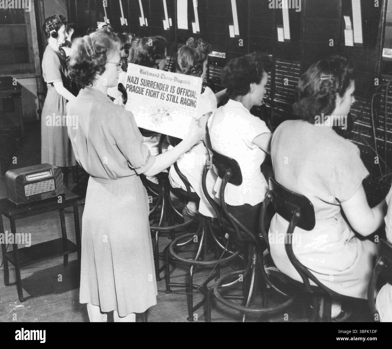 Am V-E Day geht die Arbeit wie gewohnt an der Telefonzentrale, Gebäude Nr. 201, HRPE, Newport News, Virginia. Die stellvertretende Chefmanagerin, Mrs. Bessie Fowlkes, Petersburg, Virginia, sieht sich die Zeitung an, die den Sieg ankündigt. Die Chefmanagerin, Mrs. Hanna Byrd, Washington, D.C., beaufsichtigt die Vermittler, im Hintergrund. Stockfoto